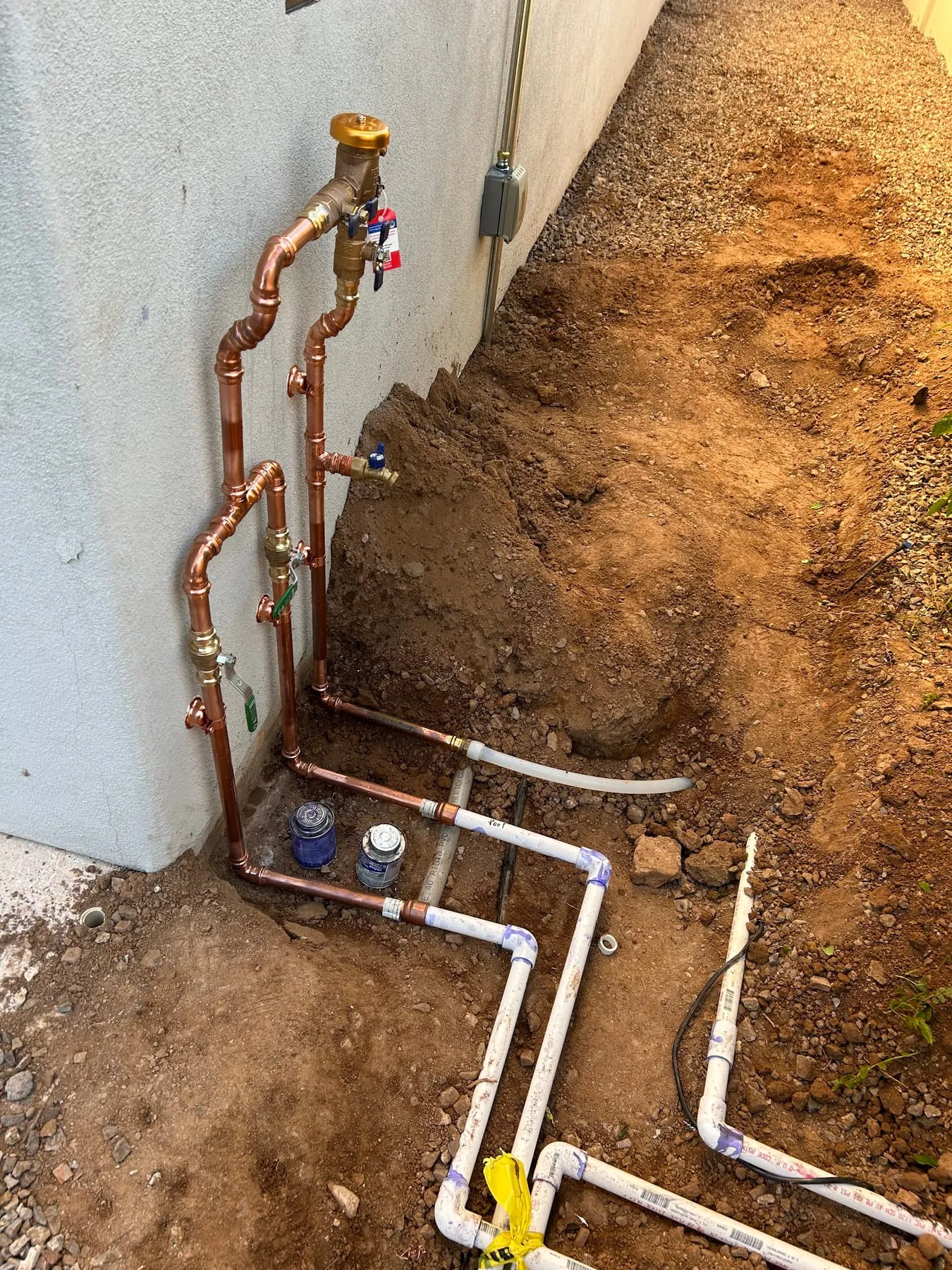 Copper supply plumbing and a brass backflow preventer on a house wall, with new PVC irrigation lines in an open trench