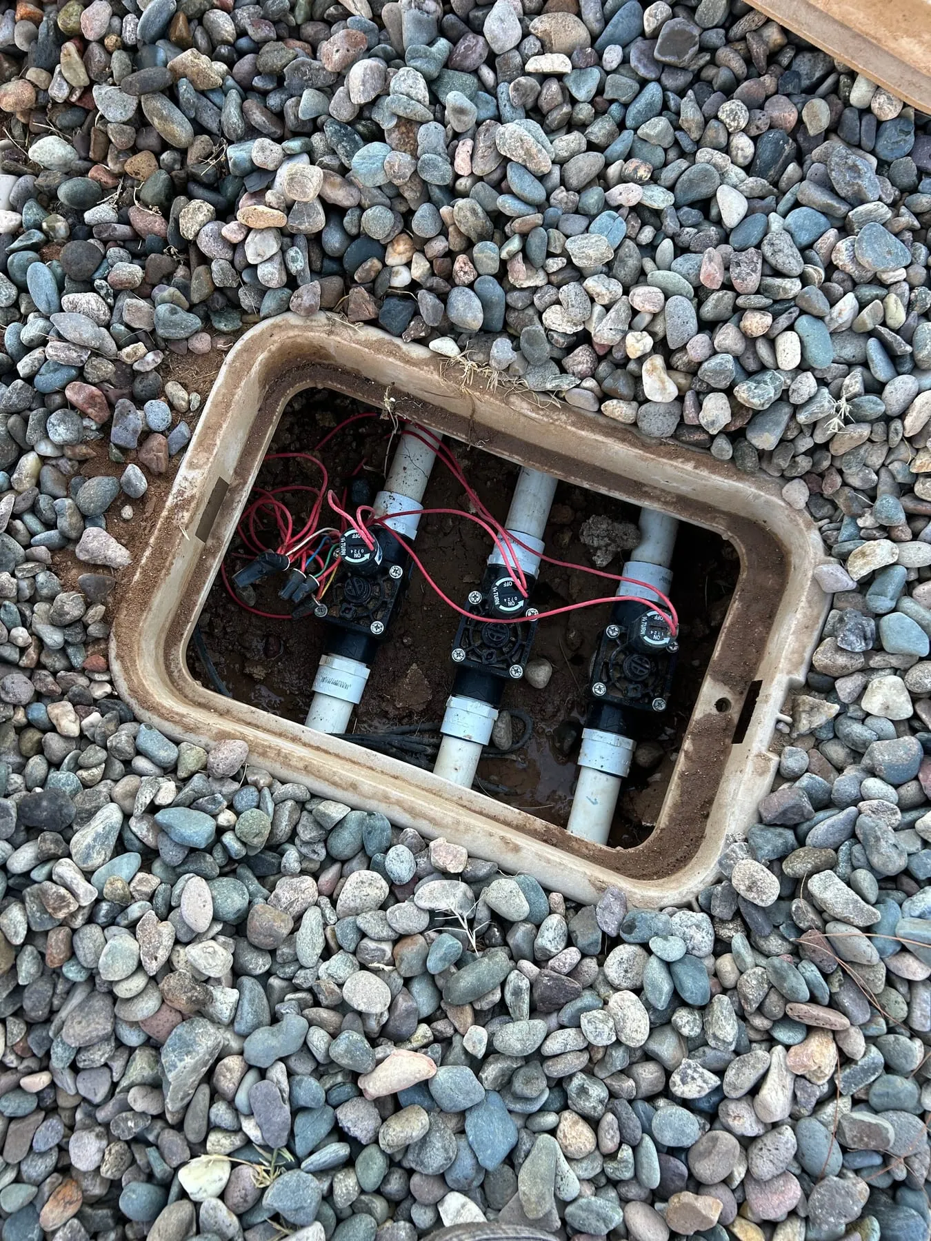 Open valve box set in river rock, showing three wired irrigation valves